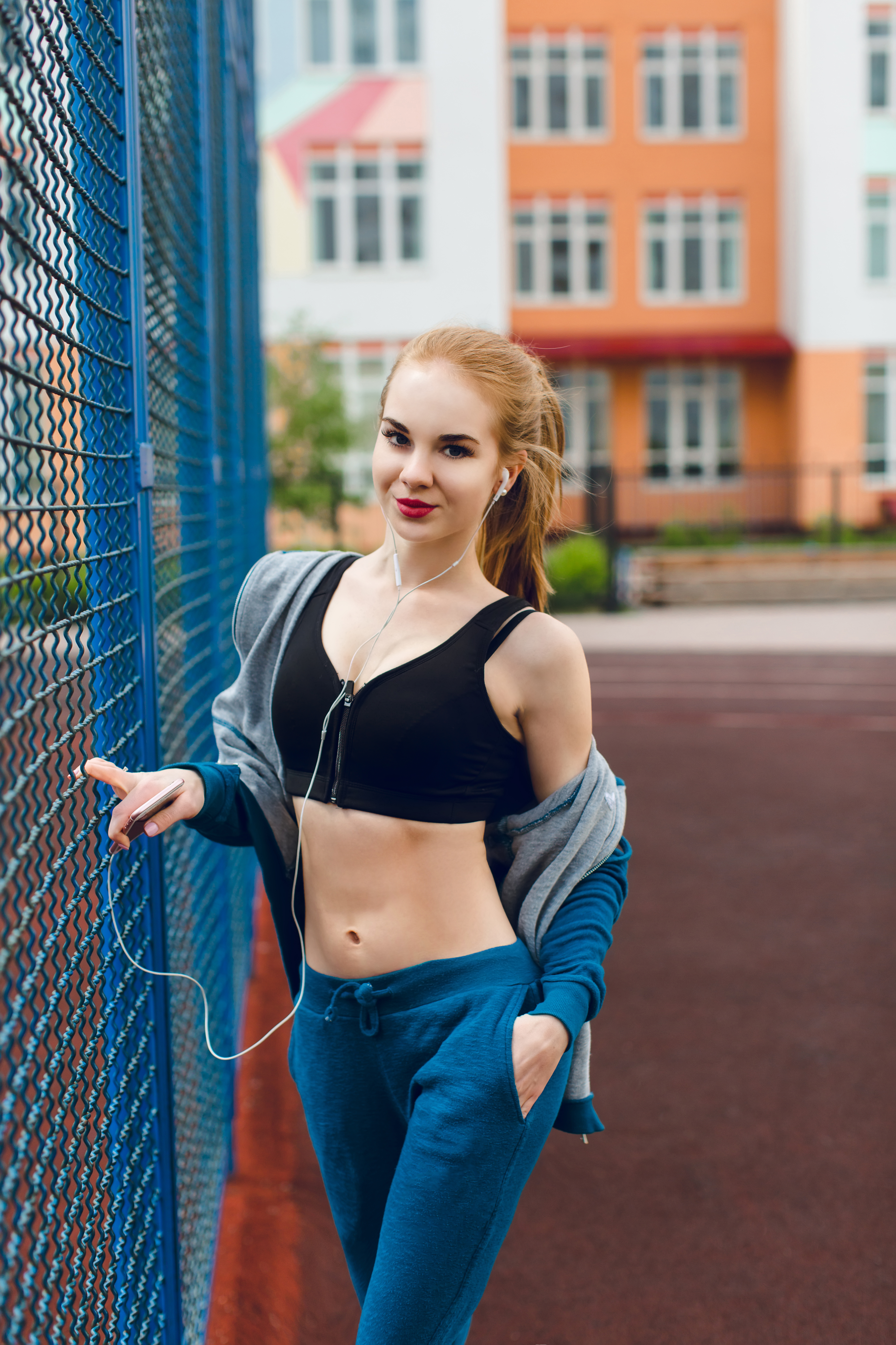 young-girl-with-good-figure-is-standing-near-blue-fence-stadium-she-wears-blue-sport-suit-with-black-top-she-is-listening-music-with-headphones-looking-camera (1).jpg